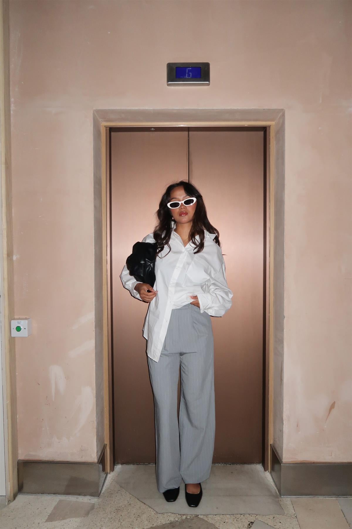 A woman standing in front of an elevator, wearing grey pinstripe wide-leg straight trousers with a high-waisted design, paired with a white top and black shoes.
