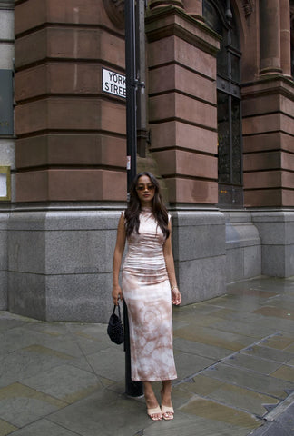 A woman standing on a sidewalk wearing a sleeveless bodycon midi-dress with a tie-dye pattern in shades of sepia.